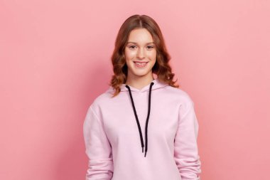 Kapüşonlu neşeli genç kadın portresi kameraya samimi bir şekilde gülümsüyor, pozitif ruh hali. Pembe arka planda izole edilmiş kapalı stüdyo görüntüleri.