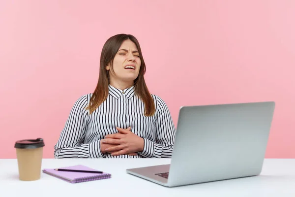 Unhappy woman office worker grimacing sitting at workplace, feeling discomfort and abdominal cramps, suffering heartburn or dysmenorrhea. Indoor studio shot isolated on pink background