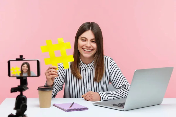 Smiling attractive woman blogger showing big yellow hashtag symbol at smartphone camera, sharing viral content, tagged message. Indoor studio shot isolated on pink background