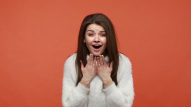 Kahverengi saçlı, tüylü beyaz kazaklı, mutlu ve neşeli bir kadın piyango zaferini ve başarısını kutluyor. Turuncu arka planda kapalı stüdyo çekimleri