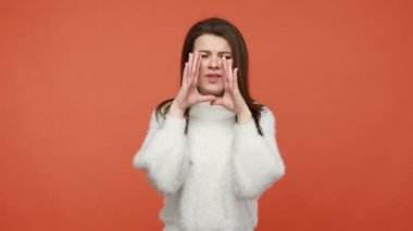 Gergin, esmer, beyaz kazaklı bir kadın ağzı açık bir şekilde el ele tutuşarak bağırıyor, negatif duygular yayıyor. Turuncu arka planda kapalı stüdyo çekimleri