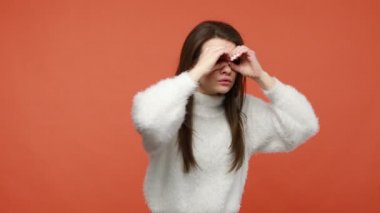 Beyaz pofuduk süveter giyen kaygısız mutlu kız parmak deliklerinden bakıyor, dürbünle el hareketi yapıyor, gözlük yapıyor ve heyecanla izliyor. Turuncu arka planda kapalı stüdyo çekimleri