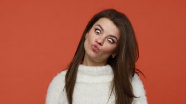 Funny brunette woman in white sweater crossing eyes making silly dumb face, having fun grimacing demonstrating dumb ridiculous face expressions. Indoor studio shot isolated on orange background