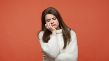 Beyaz pofuduk kazaklı sıkıcı esmer kadın kameraya ilgisiz, bitkin, ilgisiz ve tembelce bakıyor. Turuncu arka planda kapalı stüdyo çekimleri