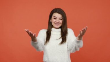 Tüylü beyaz kazaklı, dost canlısı esmer kadın. Ellerini kocaman açıyor. İçtenlikle selamlıyor ve misafirleri selamlıyor. Turuncu arka planda kapalı stüdyo çekimleri