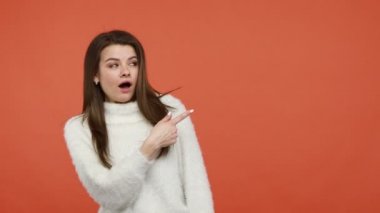 Kabarık beyaz kazaklı, boş arka plana bakan, reklam alanına, serbest alana hayretle bakan aşırı derecede şaşırmış bir kadın. Turuncu arka planda kapalı stüdyo çekimleri