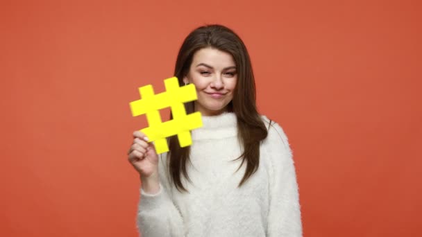 穿着蓬松的白色毛衣的积极乐观的黑发女性博主或自由职业者拿着黄色的Hashtag标牌 在网站上标注单词 在橙色背景下被隔离的室内拍摄 — 图库视频影像