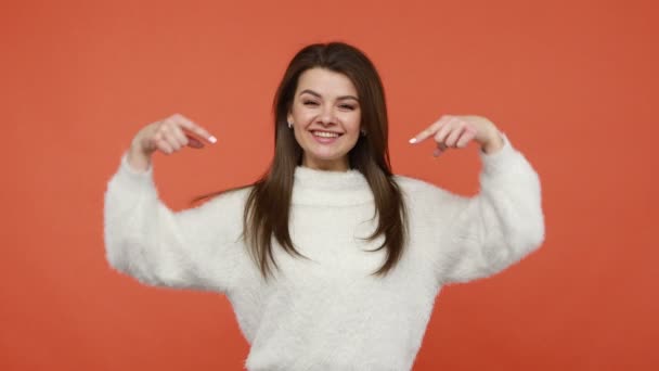 広告エリアで指を指差して親指を立てるふわふわの白いセーターに肯定的な満足した女性は 喜んでカメラを見て笑っています オレンジの背景に隔離された屋内スタジオショット — ストック動画