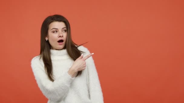 広告エリア 空き領域での驚きを探して 空の背景に脇を指してふわふわの白いセーターで非常にショックを受けた女性 オレンジの背景に隔離された屋内スタジオショット — ストック動画