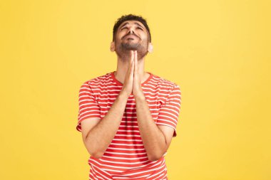 Çizgili tişörtlü, sakallı, çaresiz bir adam ellerini kavuşturup yukarı bakıyor, Tanrı 'yla iletişim kuruyor, af ve kutsama diliyor. Sarı arka planda kapalı stüdyo çekimleri