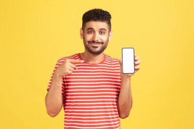 Çizgili tişörtlü, sakallı, beyaz ekranlı akıllı telefonu işaret eden, uygulamayla övünen, kendine güvenen olumlu bir adam. Sarı arka planda kapalı stüdyo çekimleri