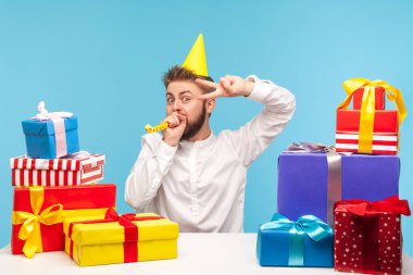 Külahta şapkalı mutlu bir adam parti üfleyicisinde uçuyor ve iş yerinde renkli hediye kutularıyla, tatil acentesiyle zafer jesti yapıyor. Mavi arka planda kapalı stüdyo çekimi