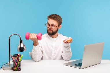 Ciddi sakallı, gözlüklü, beyaz gömlekli, bir elinde dambıl, diğerinde tatlı çörek olan bir ofis çalışanı spor yapmayı seçiyor. Mavi arka planda kapalı stüdyo çekimi