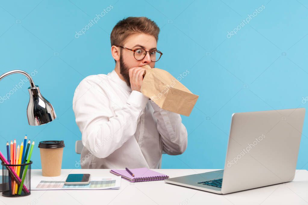 Nervous overemotional man office worker deeply inhaling and exhaling holding paper breathing bag near mouth, sitting at workplace with laptop. Indoor studio shot isolated on blue background
