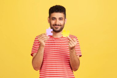Çizgili tişörtlü gülümseyen sakallı adam iki yapboz parçasını tutuyor, eşleştiriyor ve birleştiriyor, bulmacaları çözüyor, sorunları çözüyor, yaratıcılığını gösteriyor. Sarı arka planda kapalı stüdyo çekimleri