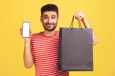 Çizgili tişörtlü, beyaz tişörtlü, elinde akıllı telefon ve siyah kese kağıdıyla gülümseyen olumlu adam online alışveriş, hızlı teslimat, pazar yeri. Sarı arka planda kapalı stüdyo çekimleri