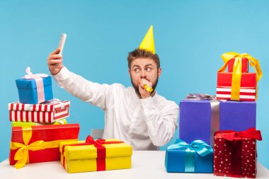 Parti şapkalı komik, pozitif sakallı adam parti kornasına üflüyor ve selfie çekiyor veya video kaydediyor, ünlü blogcu abonelere hediye hazırlıyor. Mavi arka planda kapalı stüdyo çekimi