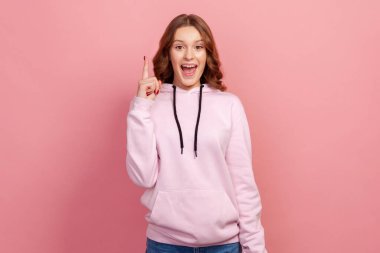 Heyecanlı, esinlenmiş, kahverengi saçlı bir gencin portresi parmağını kaldırıp kameraya dişlek bir gülümsemeyle bakıyor, yeni harika fikirle mutlu, bir çözüm buluyor. Pembe arka planda kapalı stüdyo çekimleri