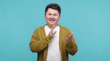 Beyaz tişörtlü ve hırkalı mutlu orta yaşlı adam yüksek sesle el çırpıyor, performansı alkışlıyor, bravo. Mavi arka planda kapalı stüdyo çekimi