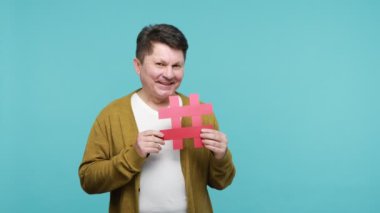 Beyaz tişörtlü ve hırkalı mutlu yetişkin adam kırmızı hashtag sembolüne bakıyor. İşaretli mesajları ve popüler virüs içeriklerini seviyor. Mavi arka planda kapalı stüdyo çekimi