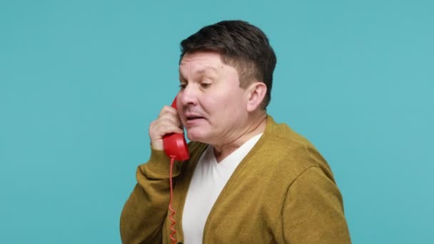 Tシャツを着た怒っている中年男性は 固定電話の電話を話している赤い携帯電話を保持叫び 不満を言い サービスセンターとの会話を保ちます 青い背景に隔離された屋内スタジオショット — ストック動画