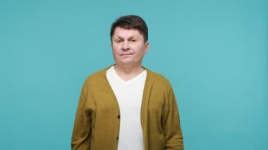 Burada ve şimdi! Otoriter, otoriter, orta yaşlı, beyaz tişörtlü ve hırkalı bir adam parmaklarını aşağıya doğru gösteriyor ve acil bir iş bitirilmesi gerekiyor. Mavi arka planda kapalı stüdyo çekimi