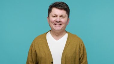 Her şey yolunda! Keyifli, orta yaşlı, siyah saçlı, tişörtlü, hırkalı, hoş bir jest yapan, dişlek tebessümle kameraya bakan bir adam. Mavi arka planda kapalı stüdyo çekimi