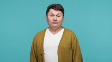 Annoyed disgruntled adult man in white t-shirt and cardigan twirling with finger near temple showing stupid gesture, feeling dumb, having insane ideas. Indoor studio shot isolated on blue background