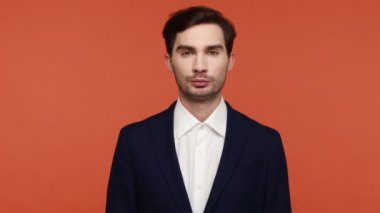 Tıraşsız, resmi kıyafet giymiş genç çekici bir adam doğrudan kameraya bakıyor ve dişleriyle gülümsüyor, olumlu duygular ifade ediyor. Turuncu arka planda kapalı stüdyo çekimleri.