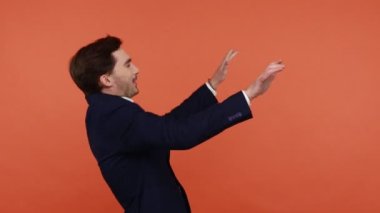 Siyah takım elbiseli pozitif sakallı adam dans ederek ve ritim tutarak eğleniyor, olumlu duygularını ifade ediyor, kutlama yapıyor ve kafayı buluyor. Turuncu arka planda kapalı stüdyo çekimleri