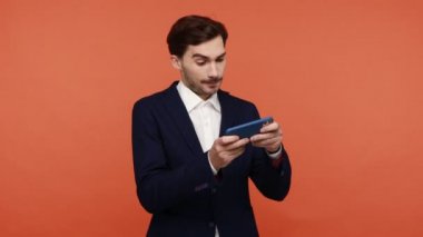Sakallı ofis çalışanı eğleniyor, tatilde akıllı telefonda sanal oyunlar oynuyor, kazanırken mutluluğu hissediyor, yumruğunu sıkıyor, alet bağımlısı oluyor. Turuncu arka planda kapalı stüdyo çekimleri