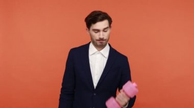 Resmi takım elbise giyen sakallı yakışıklı bir adam işte mola verirken dambıllarla egzersiz yapıyor, kaslarıyla gurur duyarak vücudunu formda tutuyor. Turuncu arka planda kapalı stüdyo çekimleri