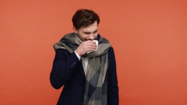 Siyah takım elbiseli, atkıya sarılmış, ağzı mendille kaplanmış, burnu akıyor ve öksürüyor, hapları gösteriyor. Turuncu arka planda kapalı stüdyo çekimleri