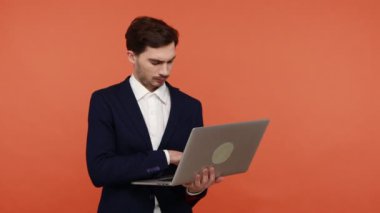 Dizüstü bilgisayarla çalışan dalgın sakallı bir işadamı, yeni bir proje düşünüyor, programlıyor, bilgisayar tutuyor, daktilo ediyor, son teslim tarihini yazıyor. Turuncu arka planda kapalı stüdyo çekimleri.