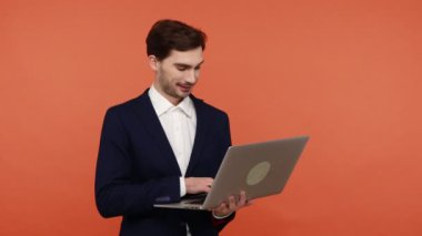 Laptopta çalışan sakallı iş adamı, klavyeyle yazı yazan, ekranda gülümseyen, işinden zevk alan. Turuncu arka planda kapalı stüdyo çekimleri