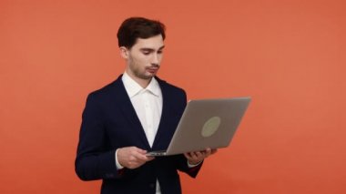 Genç, şaşkın iş adamı bilgisayarda çalışıyor ve şok edici bilgiler buluyor. Gözleriyle ekrana bakıyor, şaşırıyor. Turuncu arka planda kapalı stüdyo çekimleri