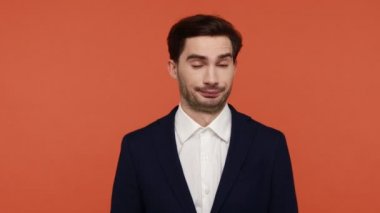 Displeased brunette bearded man in suit making stupid gesture with finger near head, looking at camera sadly, frustrated expression, dumb idea. Indoor studio shot isolated on orange background