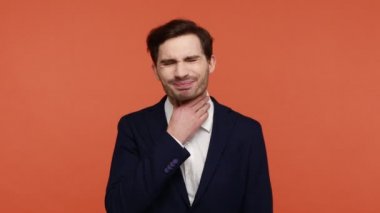 Boğaz ağrısı. Takım elbiseli genç bir adam boynunu ovuyor ve acı çekiyor boğazında ya da bademciklerinde iltihap var, sağlıksız olmak kötü hissettiriyor. Turuncu arka planda kapalı stüdyo çekimleri