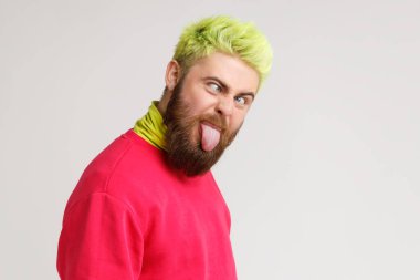 Yakışıklı, beyaz, aptal bir adamın fotoğrafı. Kameraya şaşı gözlü ve dilli bakıyor. Kırmızı kazak giyiyor, sarı saçlı. Gri arka planda izole edilmiş kapalı stüdyo görüntüleri.