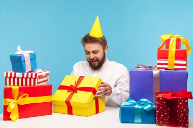 Parti şapkalı doğum günü çocuğu hediye kutusunu açıyor ve içine bakıyor, tatilin tadını çıkarıyor, bir sürü hediye alıyor. Mavi arka planda kapalı stüdyo çekimi