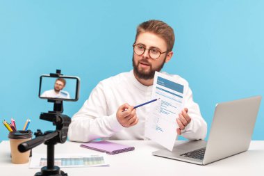 Concentrated bearded man analyst explaining how to work with data sitting at workplace at office and posing at smartphone camera, recording tutorial. Indoor studio shot isolated on blue background   clipart
