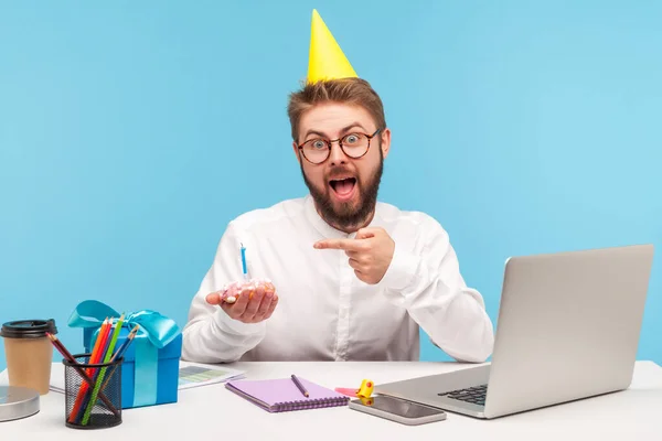 戴着眼镜 头戴派对帽 拿蜡烛指着小纸杯蛋糕的有趣而兴奋的男人 坐在工作场所庆祝他的公司周年纪念日 室内拍摄被蓝色背景隔离 — 图库照片