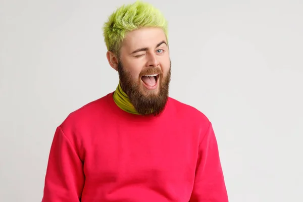 Cheerful playful young Caucasian man with beard in good mood, smiling broadly and winking at camera, keeps mouth open, dresses red jumper. Indoor studio shot isolated on gray background. — Stock Image Cheerful Playful Young Caucasian Man Beard Good Mood Smiling Broadly Stock Picture
