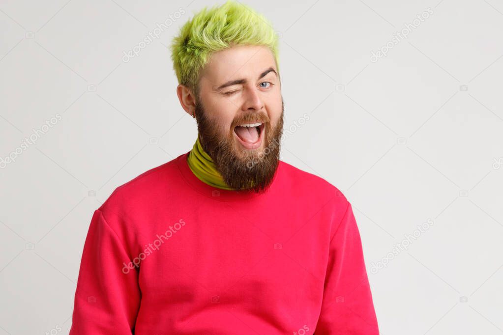 Cheerful playful young Caucasian man with beard in good mood, smiling broadly and winking at camera, keeps mouth open, dresses red jumper. Indoor studio shot isolated on gray background.