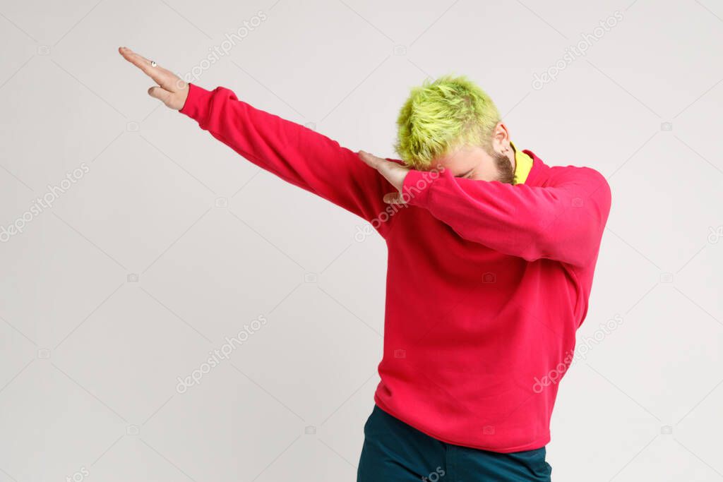 Photo of excited man with yellow hair, showing popular internet meme pose, celebrating success victory, dabbing trends, expresses positive. Indoor studio shot isolated on gray background.