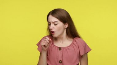 Kahverengi saçlı, öksüren, ağzı yakınında yumruğunu tutan, hasta hisseden, iltihap ya da akciğer iltihabı geçiren sağlıksız bir kadın. Sarı arka planda kapalı stüdyo çekimleri.