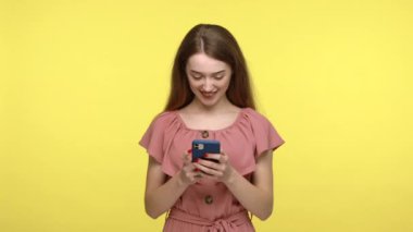 Hoş görünümlü gülümseyen mutlu kız şık telefonlarda sosyal ağ paylaşımları yapıyor, iyimserliğini dile getiriyor, cep telefonundan memnun. Sarı zemin üzerinde kapalı çekim izole