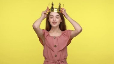 Ben en iyisiyim! Altın taç takan, kendine güvenen, kibirli, güzelliği ve başarısıyla gurur duyan, gülümseyen ve kendine hayran olan bencil, tapılası bir kadın. Sarı arka planda kapalı stüdyo çekimleri.