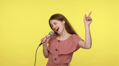 Zarif pembe elbiseli sevimli kız karaoke ve dansta mikrofonla şarkı söylüyor, neşeli bir havası var, tatlı tatlı gülümsüyor, kutluyor. Sarı arka planda kapalı stüdyo çekimleri.