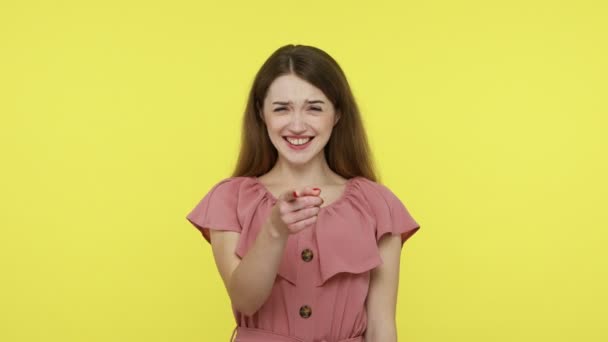 茶色の髪をした有名な女の子は カメラで指を指してピンクのドレスを着て ばかばかしい外観の楽しさを作り 大声で 陽気な笑いを笑っています 黄色の背景に隔離された屋内ショット — ストック動画